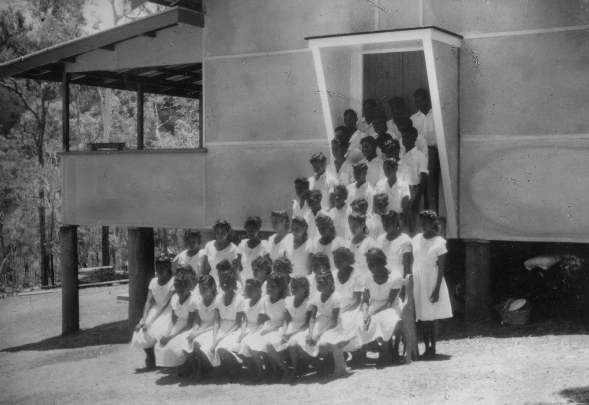 Group of young First Nations peoples wearing white clothes standing in rows in front of a building
