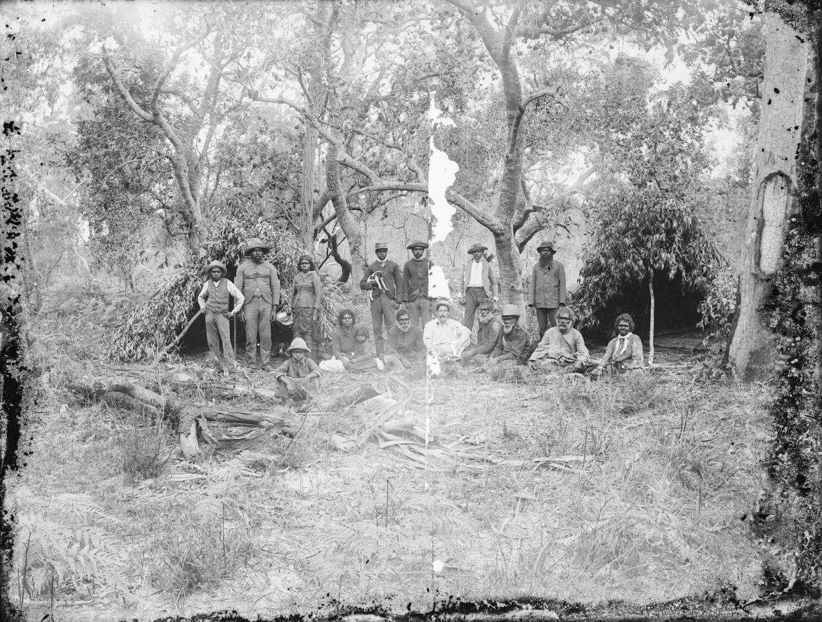 Group of First Nations people standing, and group of First Nations people and one white man sitting, posed in front of bush shelters