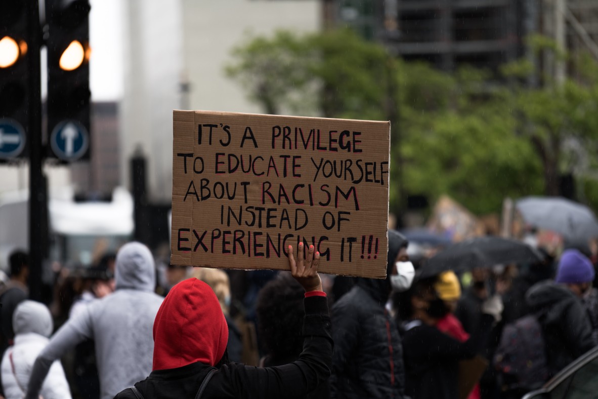 People in street rally, showing person holding up cardboard placard reading "Its a privilege to educate yourself about racism instead of experiencing it!!!"