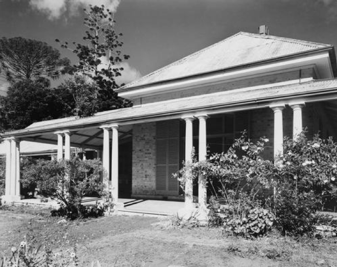 A black and white photograph of  a house with  a verandah and shrubbery around the outside