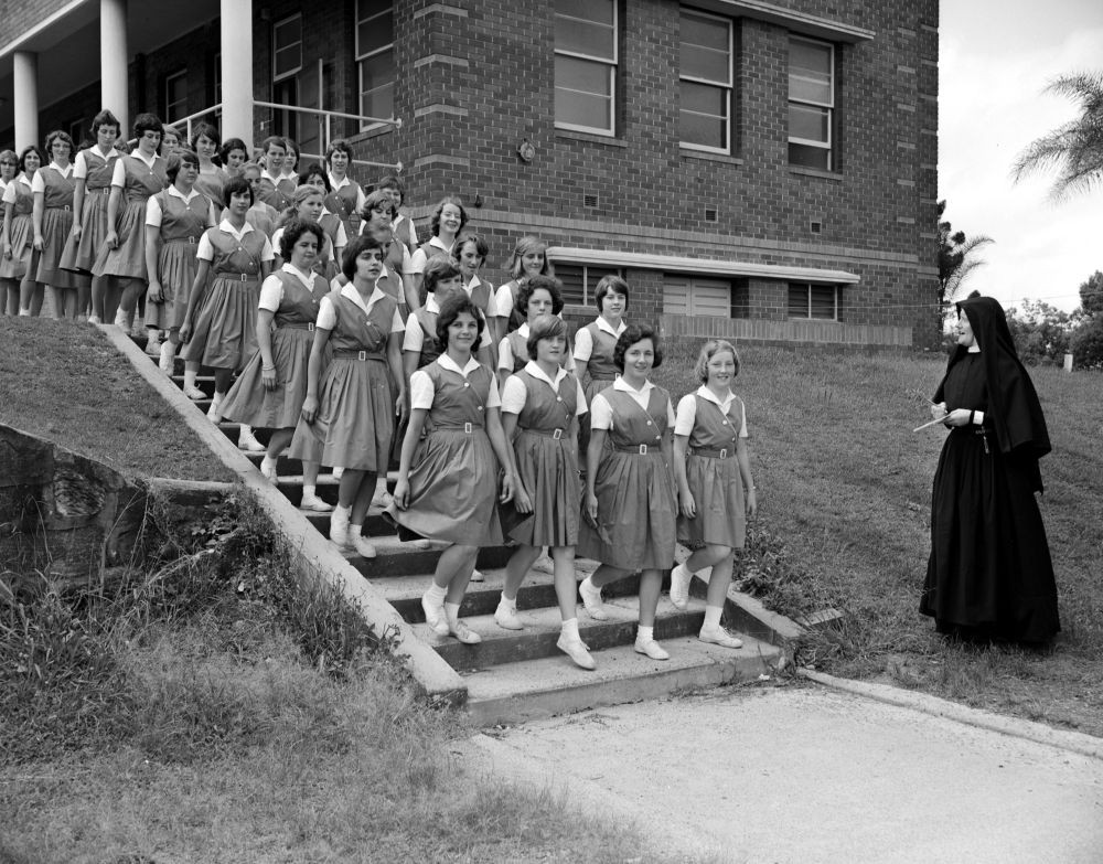 Schoolgirls descending a flight of stairs outside a school building supervised by a nun in a black habit and veil