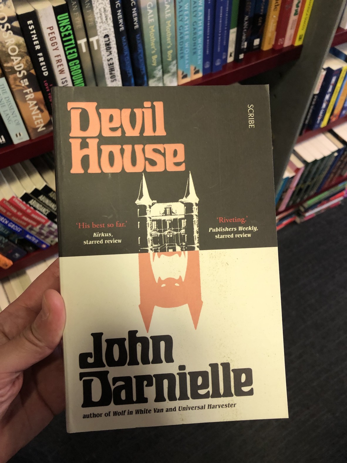 A hand is holding a copy of the book Devil House by John Darnielle In the background are bookshelves
