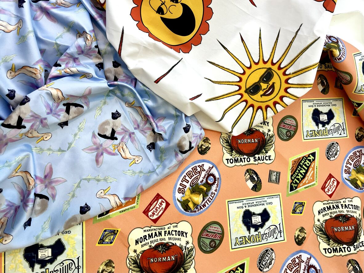 Close up photo of three different digital printed fabric on top of each other. One is white with suns, one is orange with a bunch of labels and one is blue with flowers and cats. All images used within the fabric are from State Library of Queensland collection.