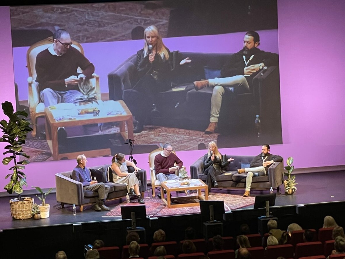 Image is of a stage at a writers festival Five people sit onstage with plants and rugs on the floor Behind them the big screen shows the speaker up close 