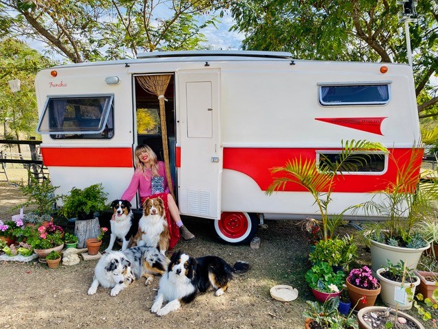 Author Holly Ringland sitting in the doorway of her red and white caravan. Four dogs are sitting in front of her and there are many potted plants to either side of the caravan.