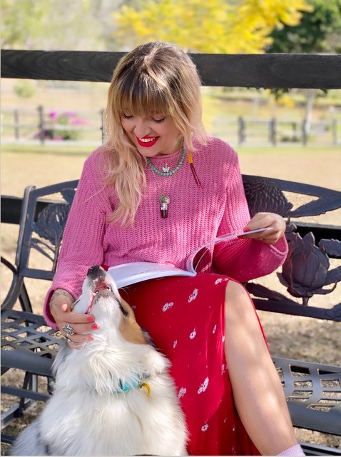 A woman sitting on an ornate metal bench with a book open in her lap. A dog sits on the ground next to her, she is smiling down at it while she pats it.