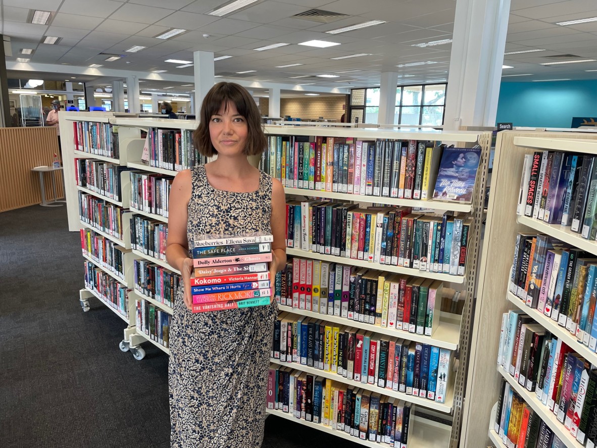 Kay holds a stack of books in Maroochydoore public library. Behind her are shelves. 