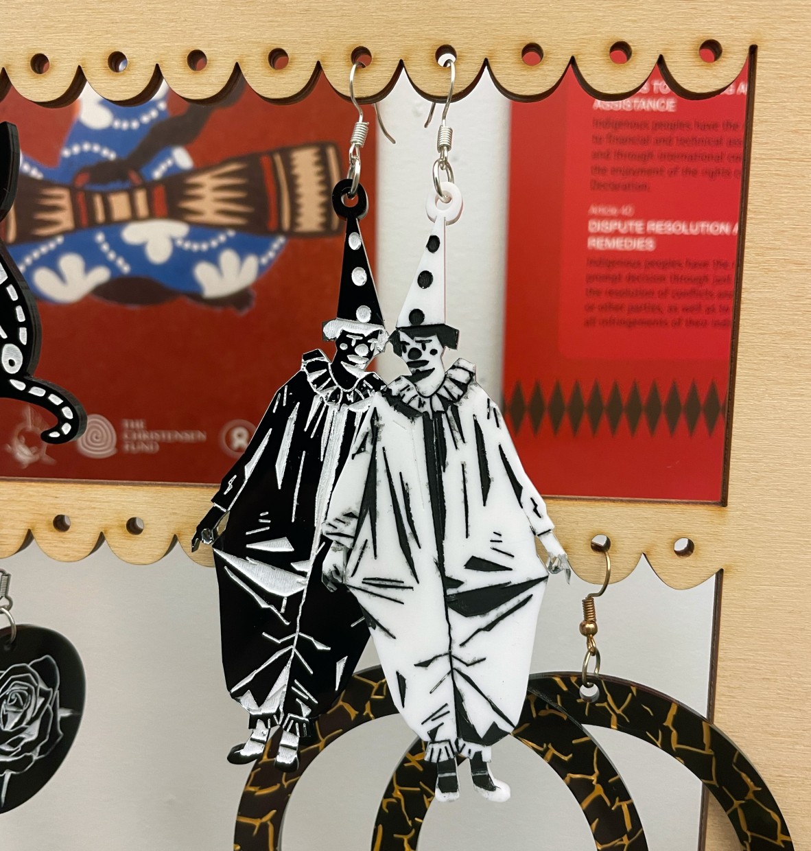 This photo shows a pair of artistic earrings featuring monochrome clown figures, one in black and the other in white. The clowns are dressed in traditional, ruffled costumes with pointed hats, giving off a vintage, somewhat eerie vibe. The earrings are displayed on a wooden holder with a scalloped edge, adding a decorative touch to the presentation.