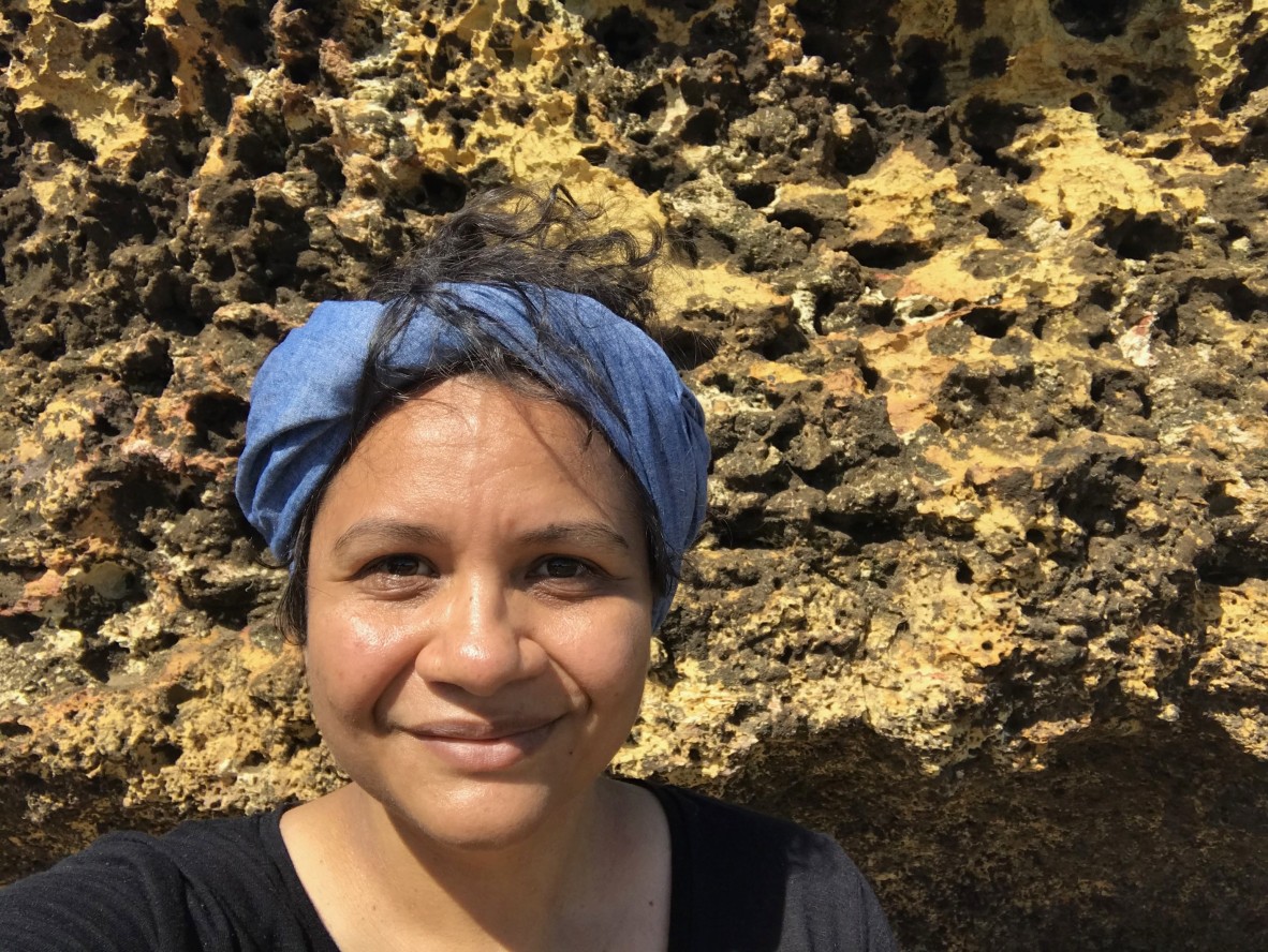 A smiling woman in a black shirt standing in front of a rock feature. She is wearing a blue bandana.