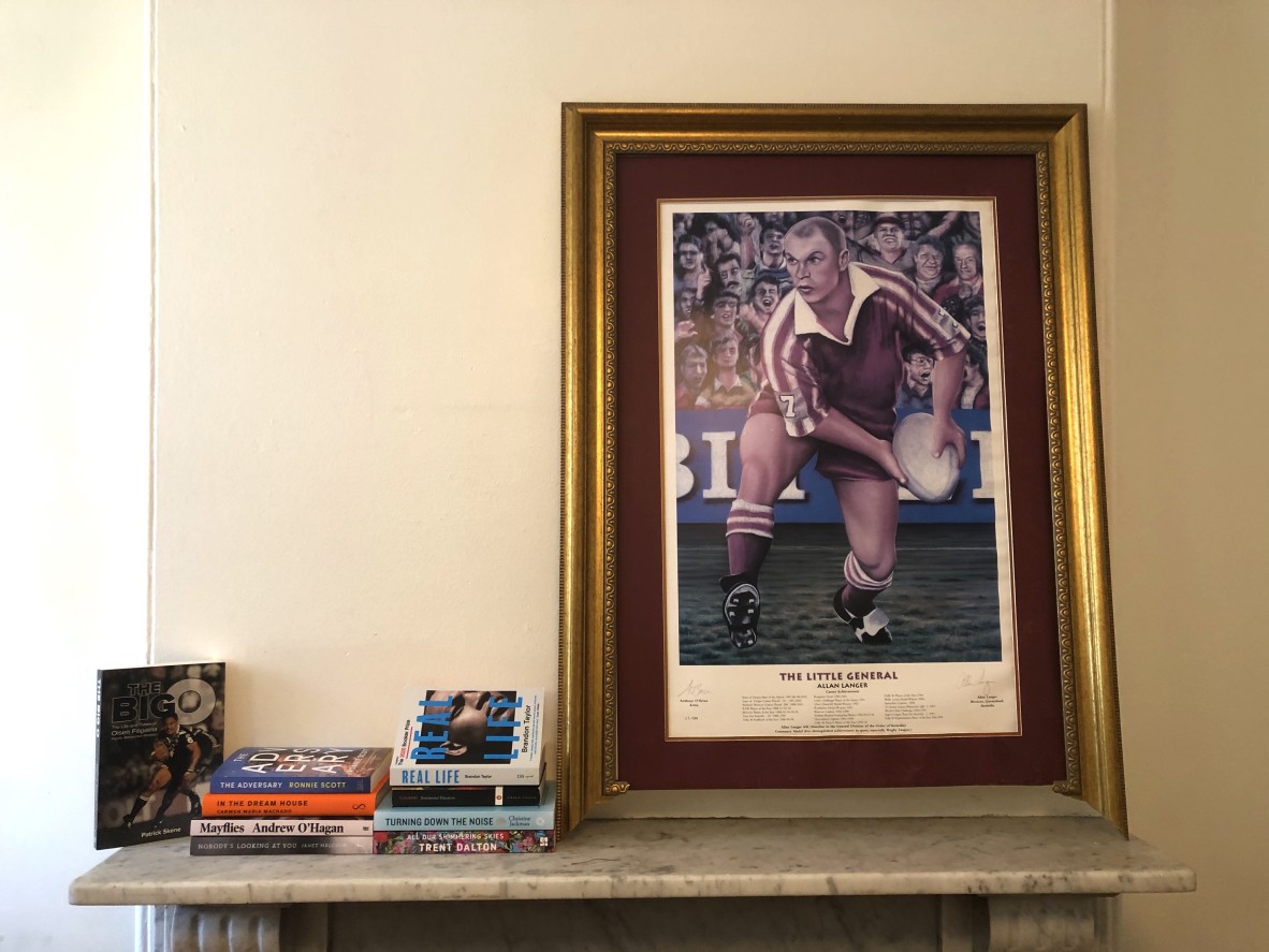 Two stacks of books sit on a mantelpiece beneath a poster of the mighty Allan Langer