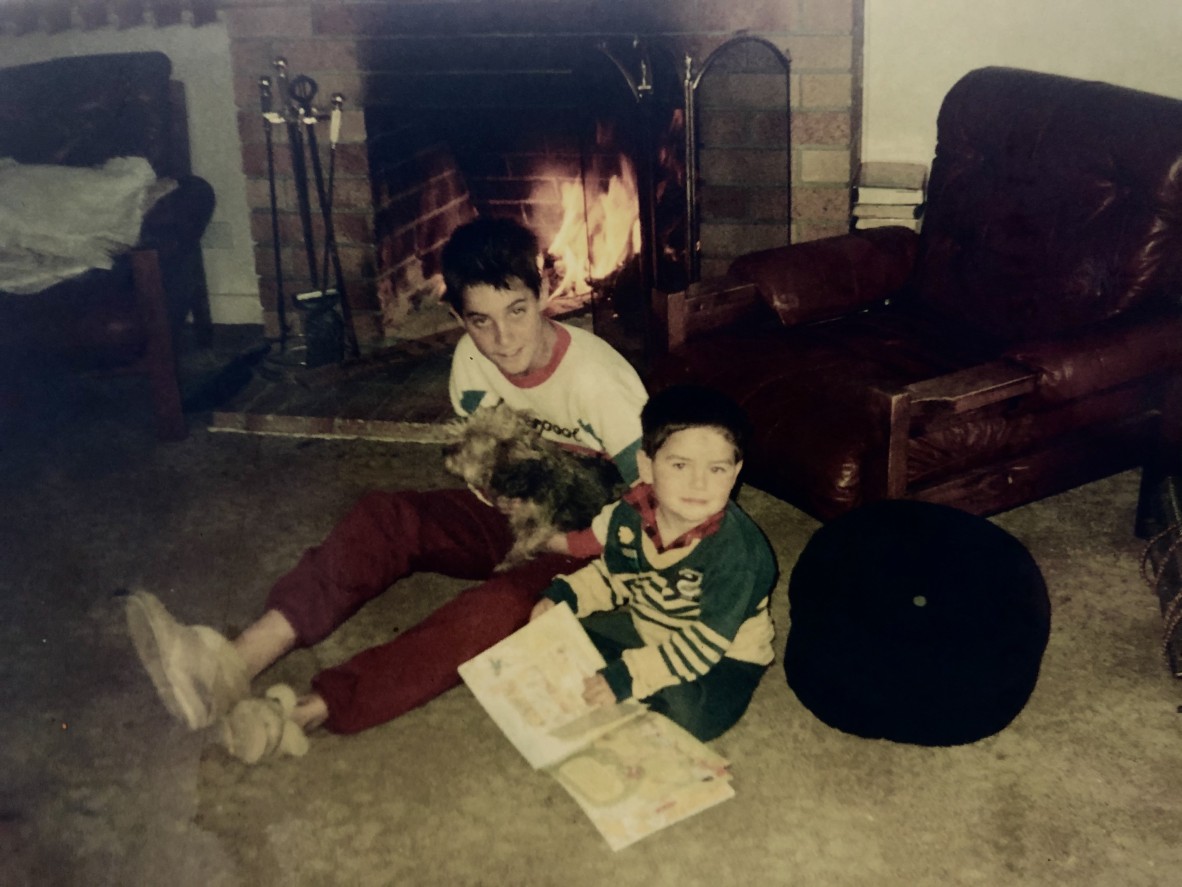 Two children read books on a lounge room floor beside a fire One of the kids is holding a dog 