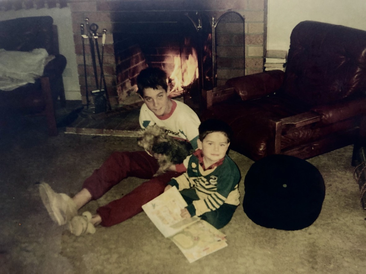Two children read books on a lounge room floor beside a fire. One of the kids is holding a dog.