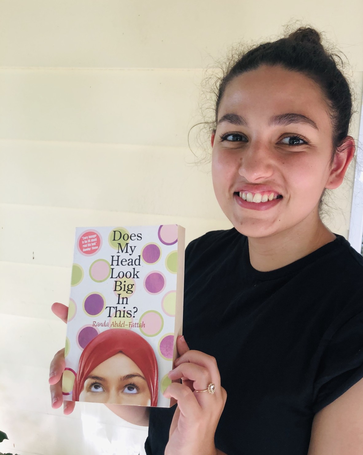Sara El Sayed wears a black t-shirt and smiles at the camera, holding a copy of the book 'Does my head look big in this?'