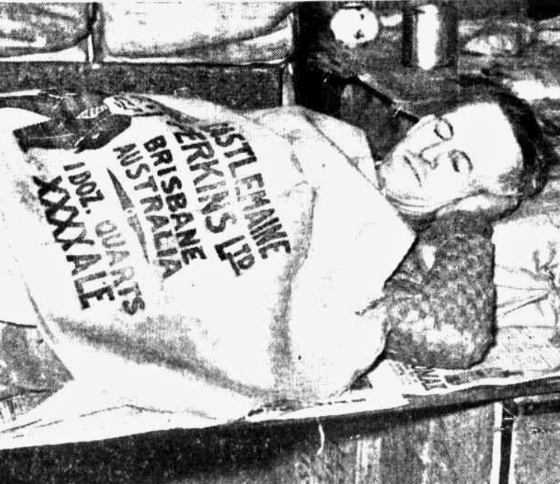 Black and white newspaper image of a man appearing asleep while covered by a sack clearly meant to contain '1 Doz. Quarts XXXX Ale'.