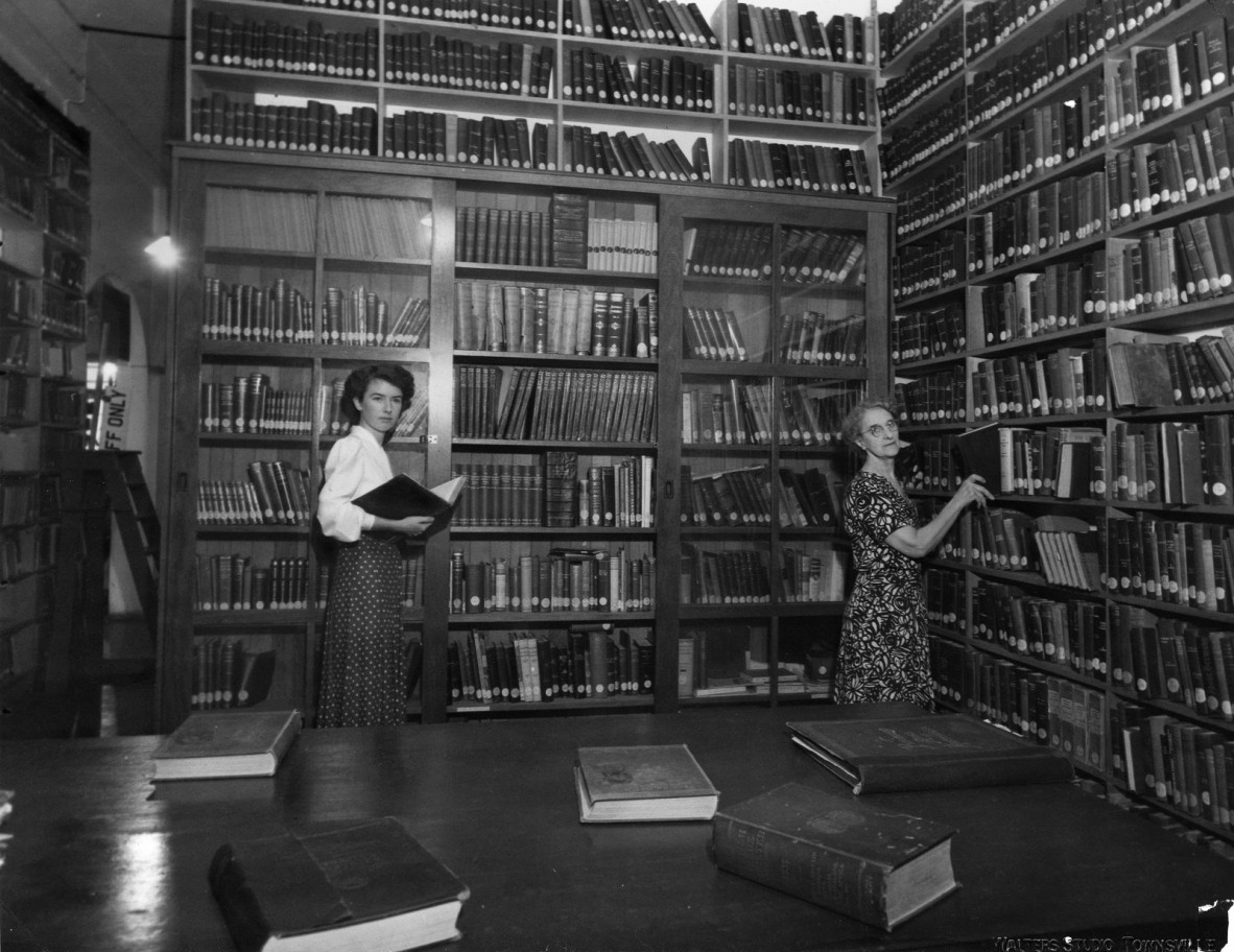  The library has floor to ceiling fixed wooden shelving with the top of a large wooden table visible in the foreground. The non-fiction section of the library with librarian Mrs Mabel C. Classen on the right. Mrs Classen held the positions of Secretary-Librarian of the Townsville School of Arts from 1921-1938, then Librarian in Charge of the Townsville Municipal Library from 1938-1964. The young lady on the left is now Mrs Meryl Stone.