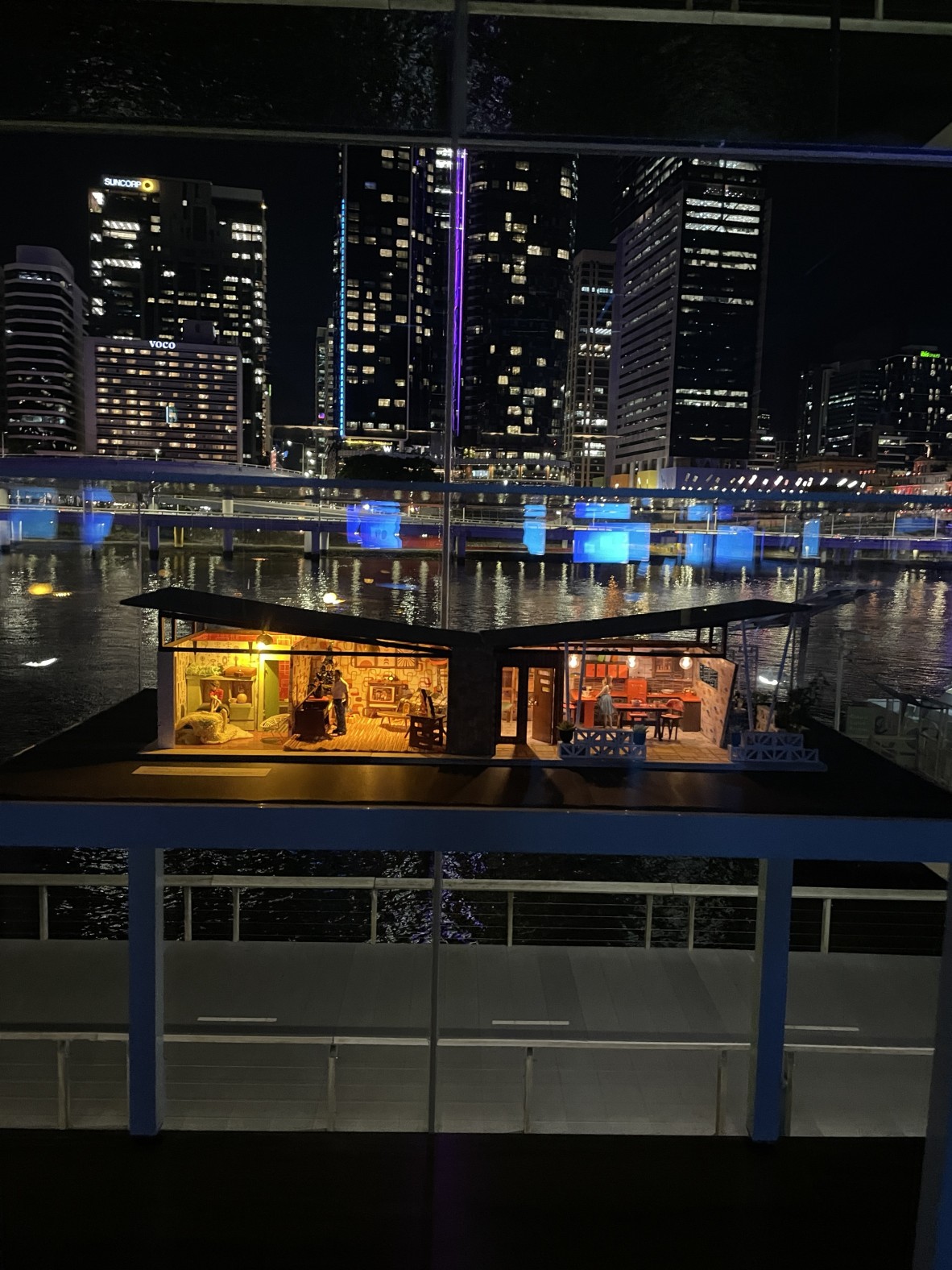 a photo at night of a mid-century model. The photo is looking at a light up model house with the Brisbane city in the background.