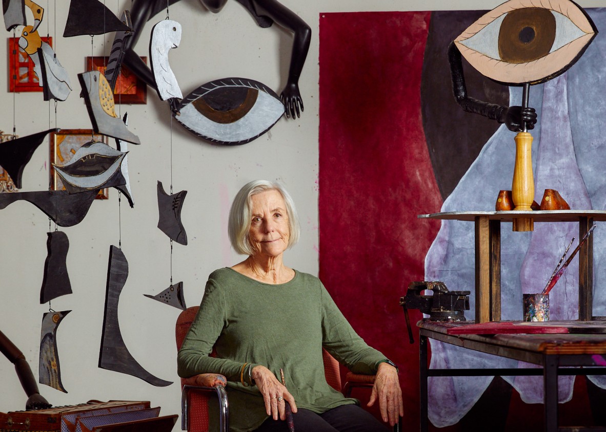 Artist Judith Wright in a green shirt sits in a chair. sculptural painted artworks hand from strings behind her, some shaped like mouths and eyes.