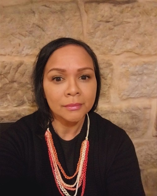 Headshot of Julie-Ann 'Garrimaa' Moore. She is seated in front of a brick wall. She wears a black top and a necklace with pink, orange and white strands