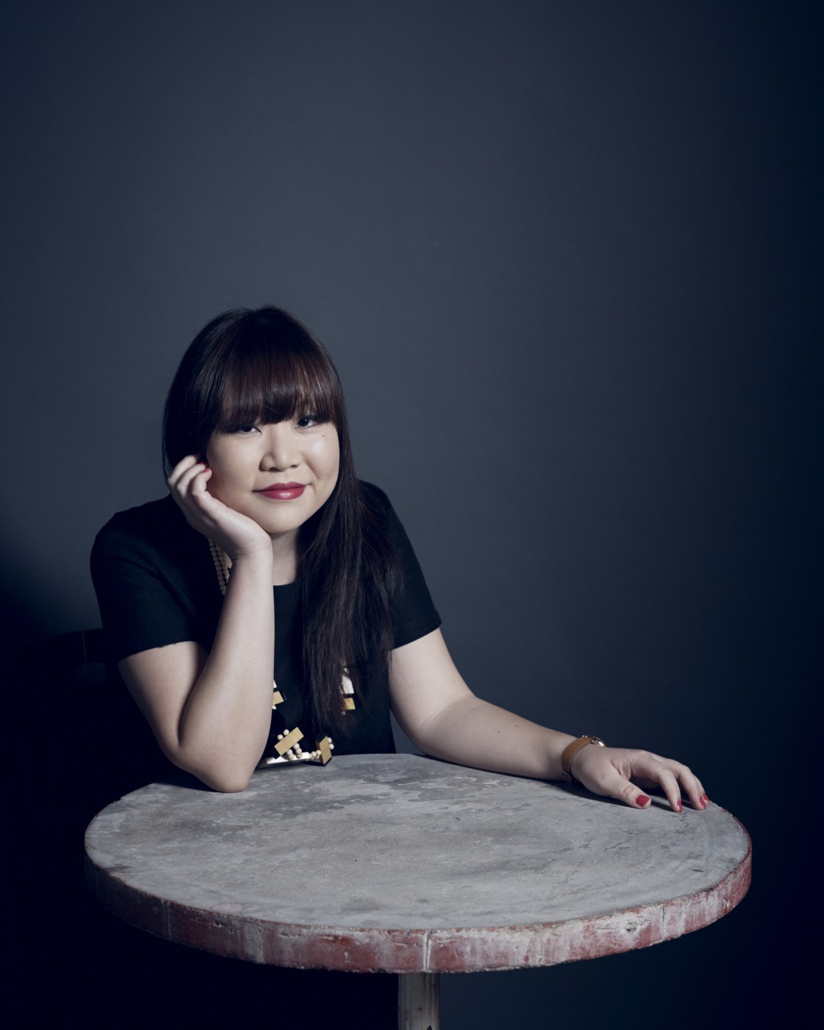 Portrait of Julie Koh sitting at a table She wears a black shirt and necklace and rests her chin in her hand 
