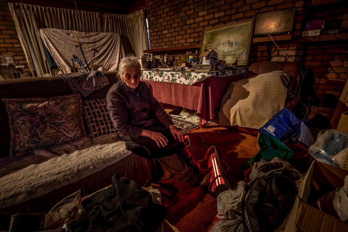 An elderly woman sits alone in a room full of furniture and items in the middle of packing