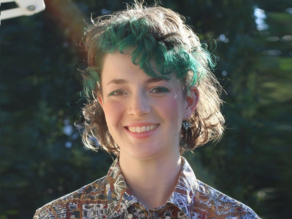 Ellie Kaddatz stands smiling outside in the sun Her hair is coloured green at the fringe and she wears a patterned shirt 