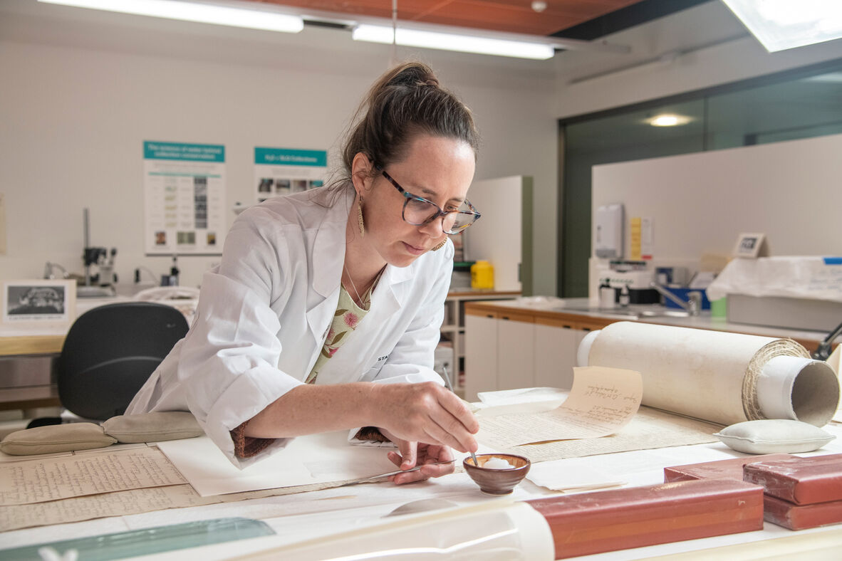 Woman in white lab coat dipping small brush into glue pot with roll of document on lab bench