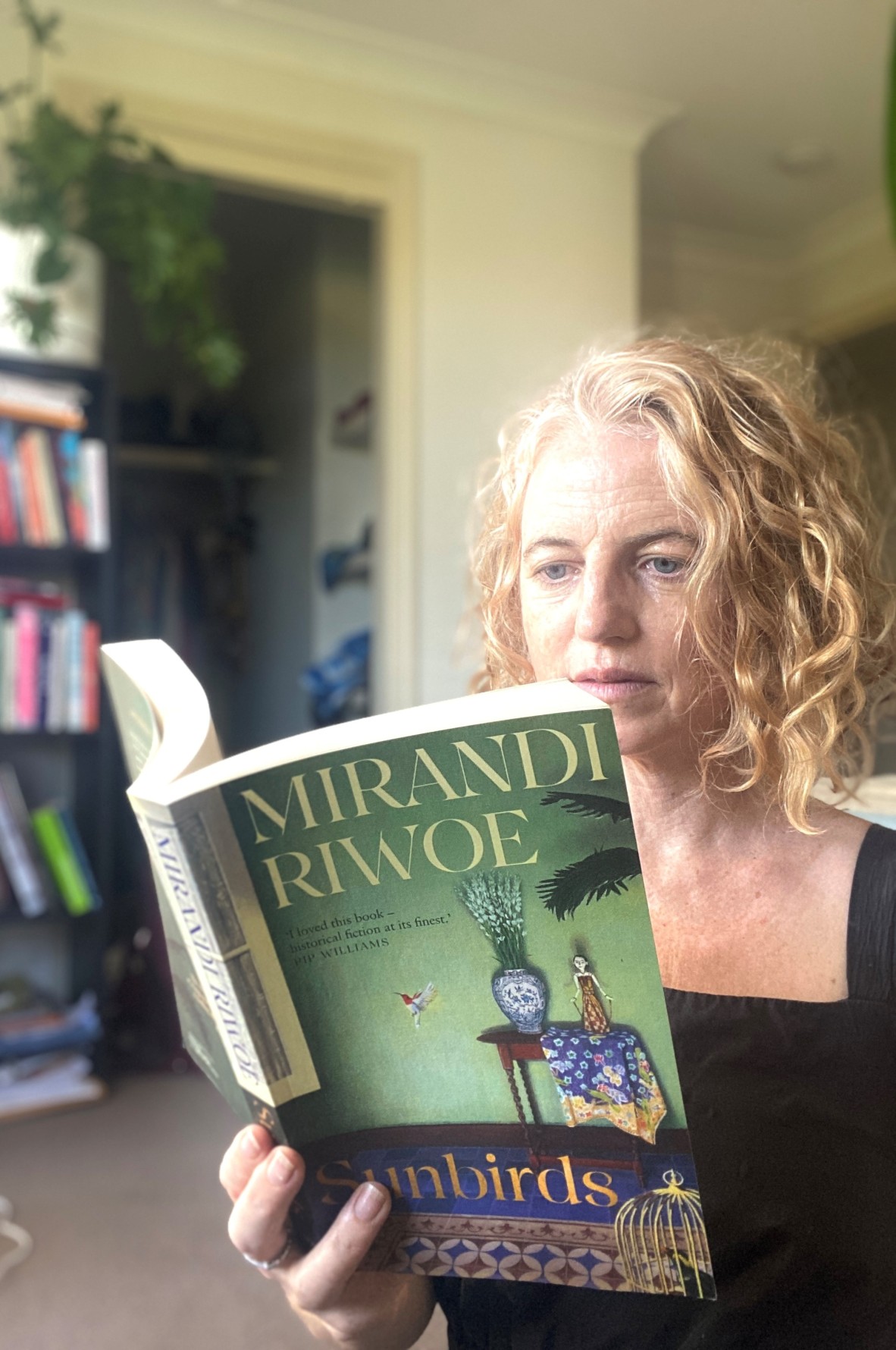Laura Jean is reading Sunbirds by Mirandi Riwoe. In the background are bookshelves and plants