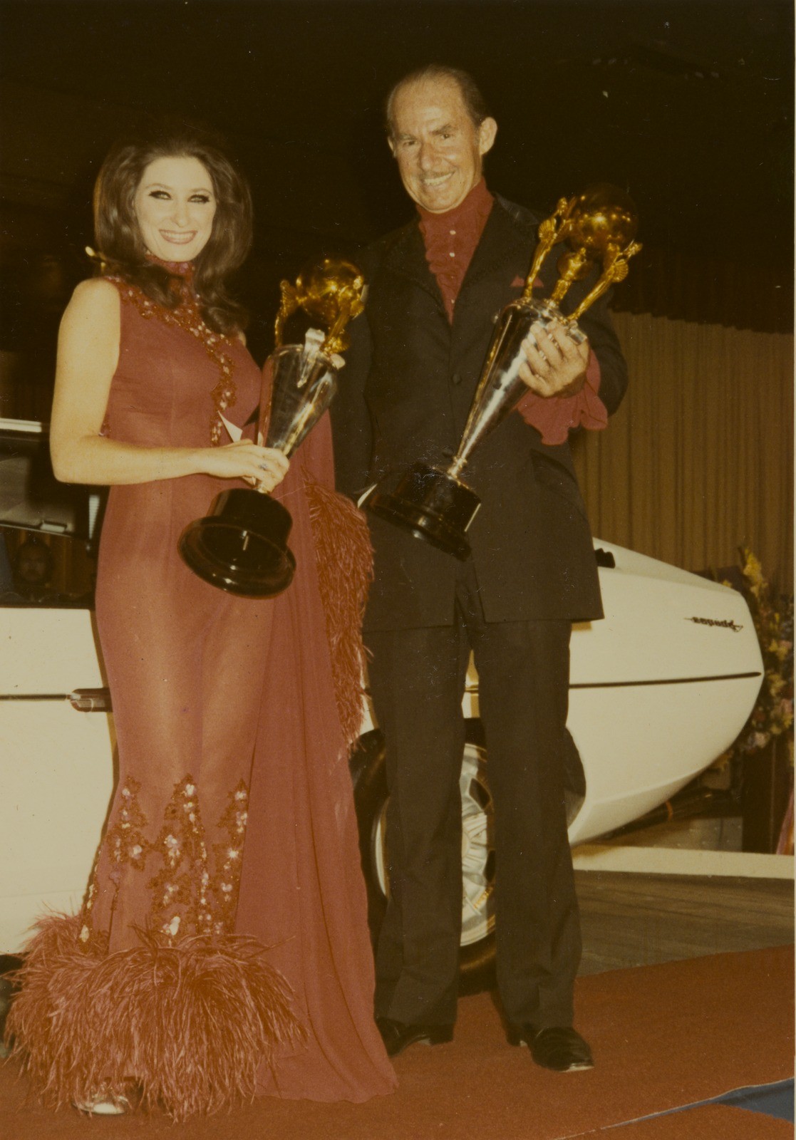 A man and woman pose holding a trophy in front of a sports car in 1971