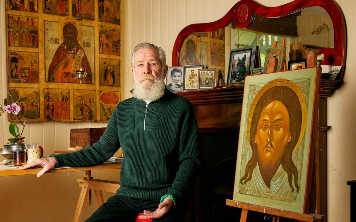 white-bearded man sitting next to icon painting displayed on easel