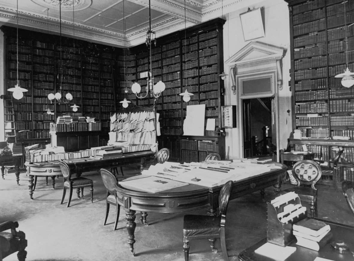 Two tables with books and newspapers on them and bookshelves around the walls in the library in Parliament House Brisbane ca 1906