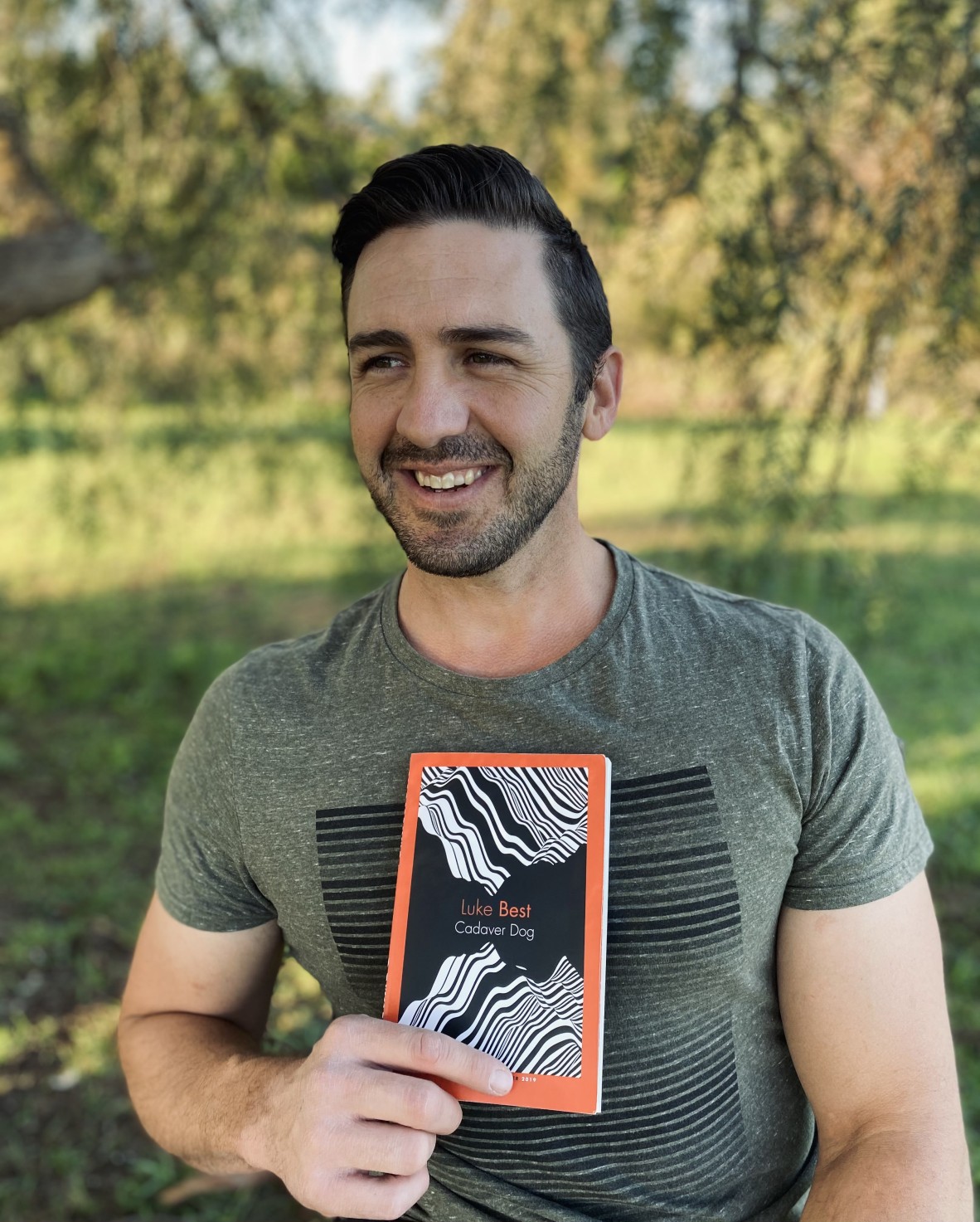 Luke Best stands beneath a tree smiling away from the camera holding his copy of Cadaver Dog