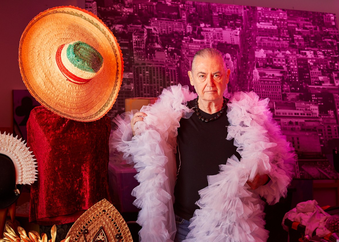 Artist Luke Roberts in a dark shirt and ruffled pink tulle coat in his studio. Complex head dresses and a sombrero can be seen to Luke's right and he stands against a pink toned background.