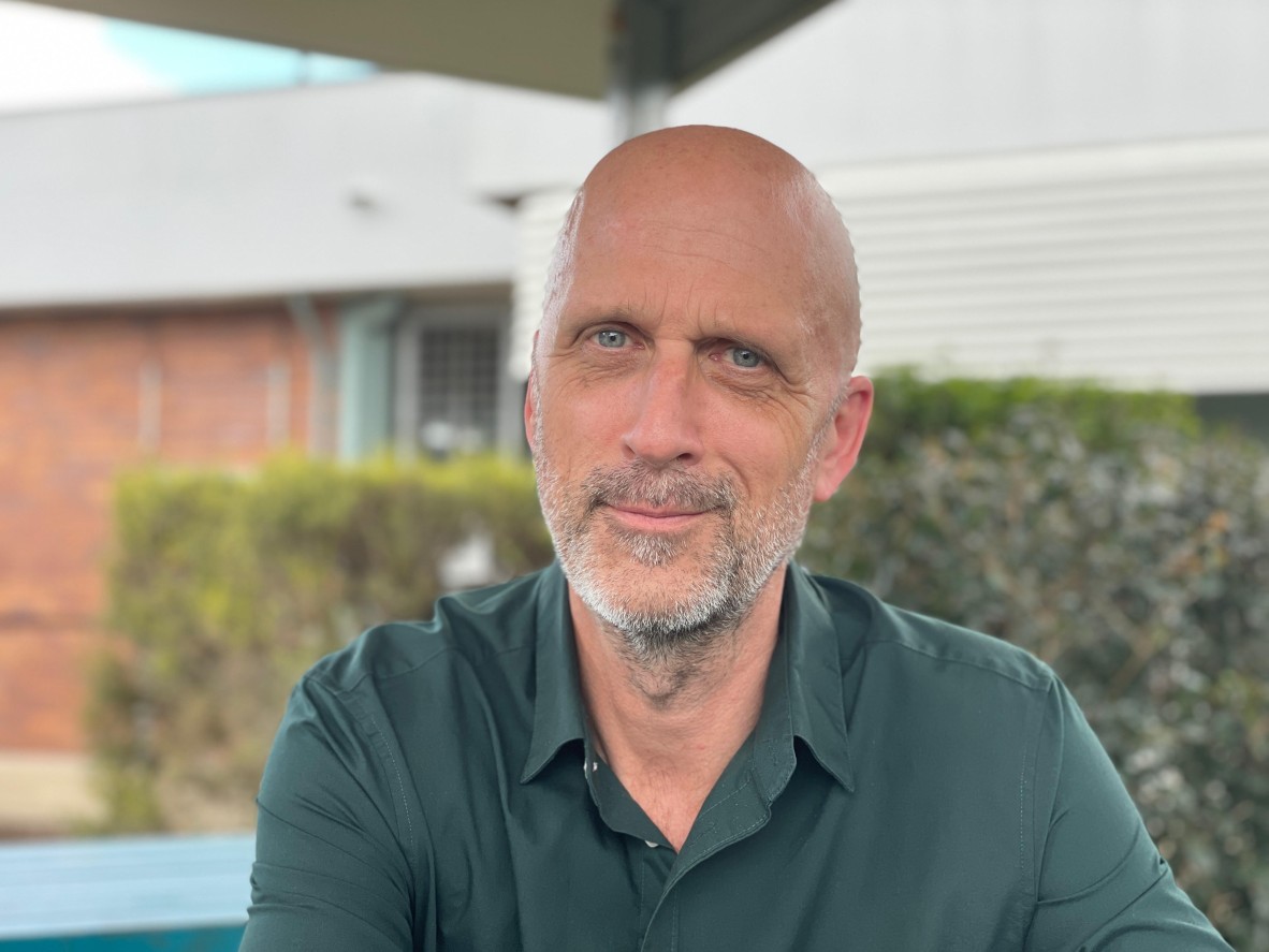 A photograph of Luke Stegemann, a writer wearing a green collared shirt, sitting outdoors in front of a brick building.