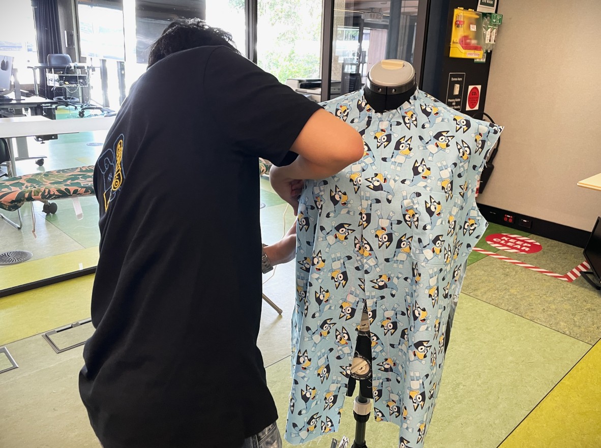 A photo of a young man next to a sewing mannequin with a shirt he is making made out of fabric featuring the character of Bluey.