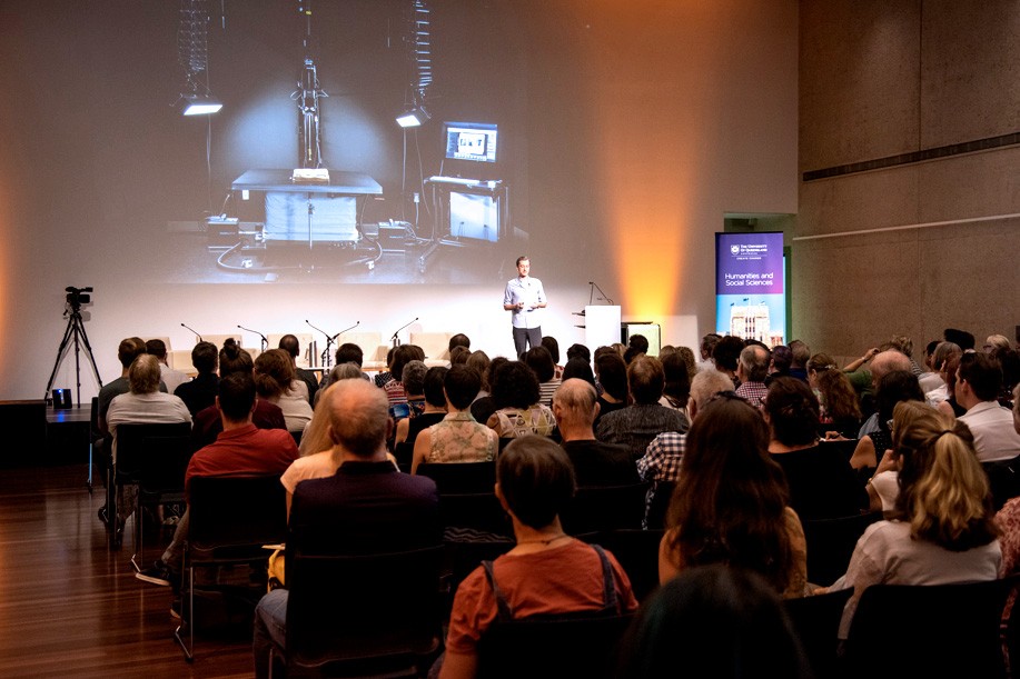 Audience seated and watching a speaker on stage with a projection screen in the background.