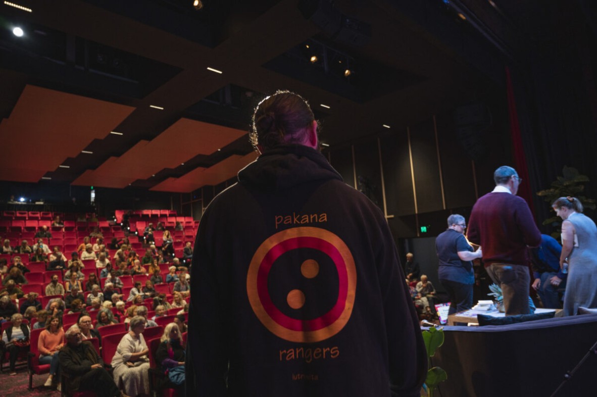 View from a stage. An audience sits on red chairs. A man's back is visible and performers are getting microphones fitted. 