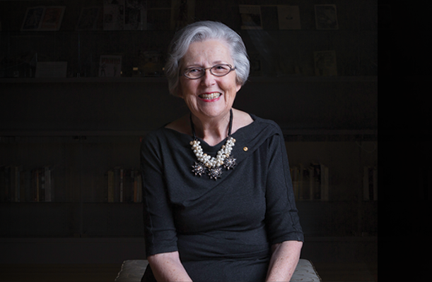 Dr Marie Siganto AM, seated in a library in front of shelves of books