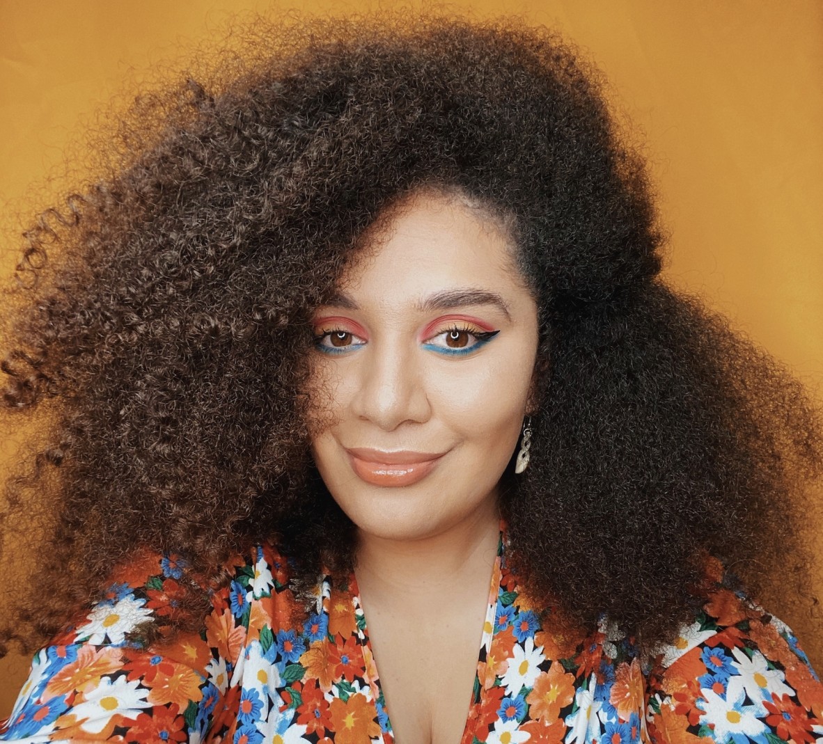 A smiling person in front of an orange background wearing a multicoloured top. Their eye makeup matches their clothes in a palette of orange and blue.