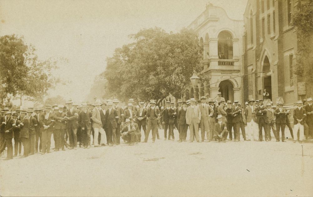 Large group of men standing gathered in Albert Square in Brisbane 1912 with two buildings behind them on the right.