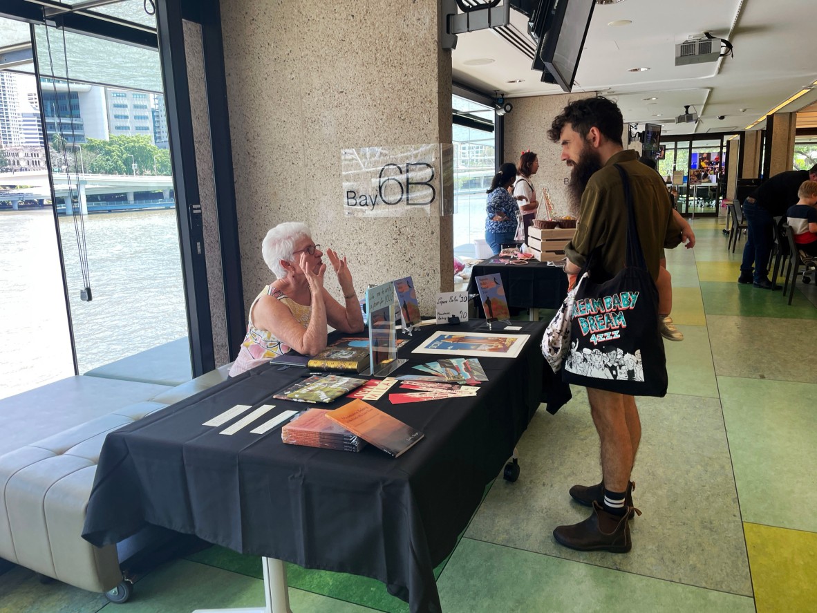 A client sitting down in front of a stall talking about their wares with someone standing in front of the table. The stall is covered in books and poetry items.