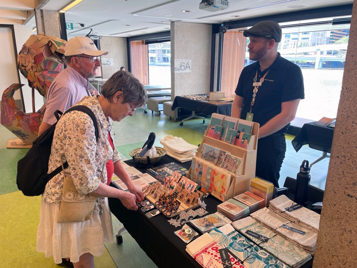 Two people looking at a market stall. The stall was run by the Library Shop and holds lots of QLD and First Nations items.