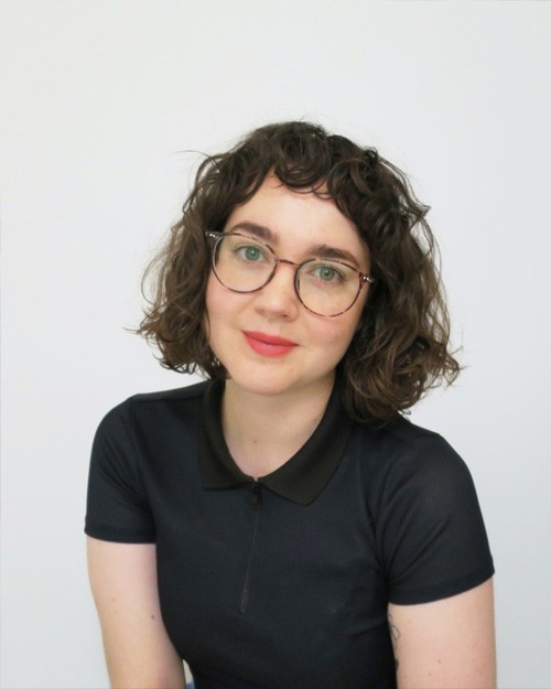 Headshot of Miranda Hine who is wearing a short sleeved black top, red lipstick and glasses. She is looking at the camera