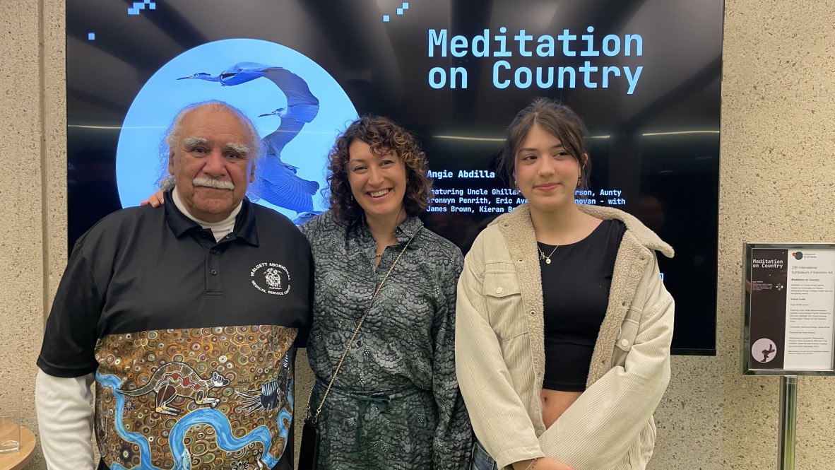 A photo of three people posing in front of a screen which has the title Meditation on Country displayed