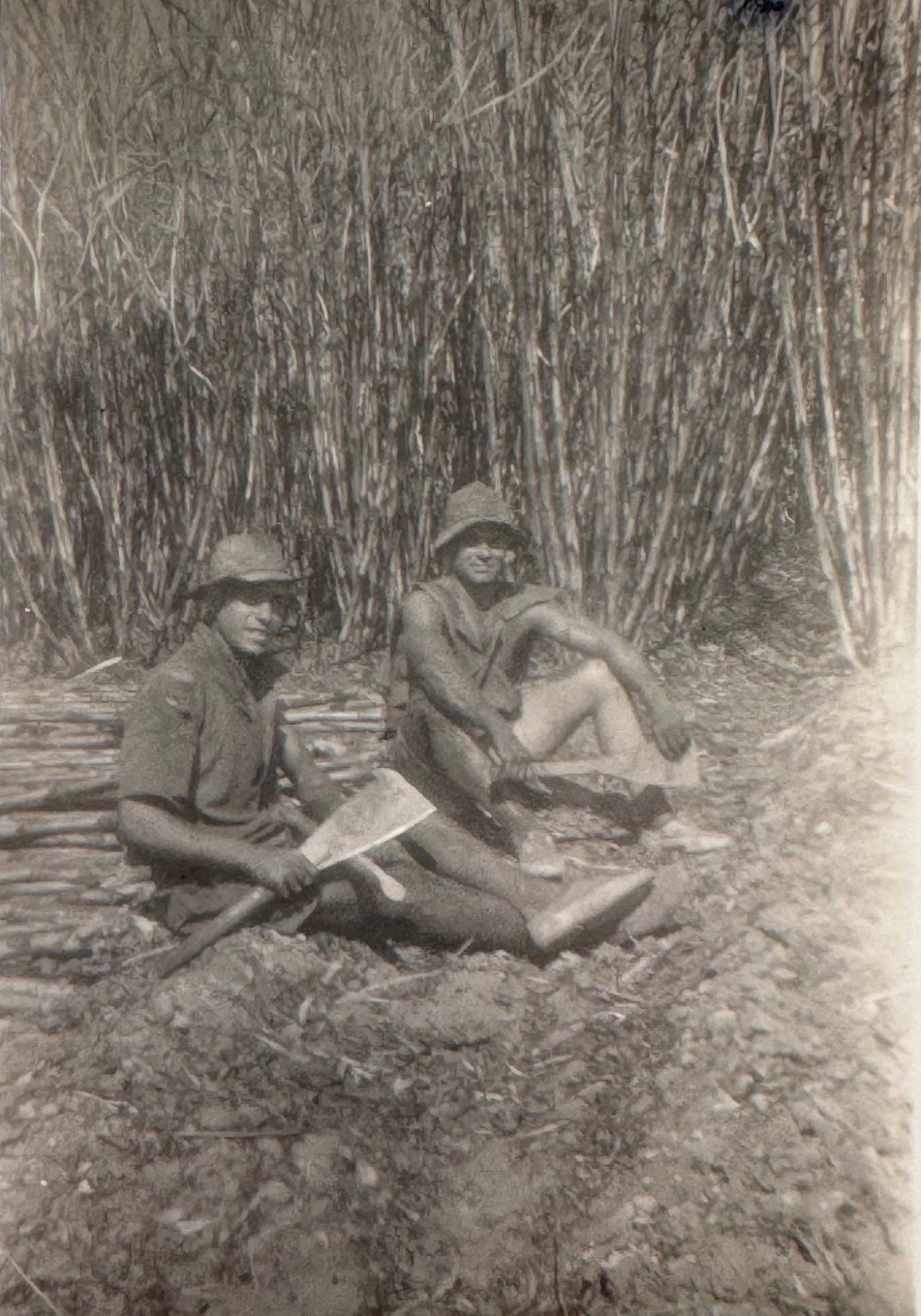 Two men sitting on ground in front of sugar cane field. They are holding cane cutting tools.