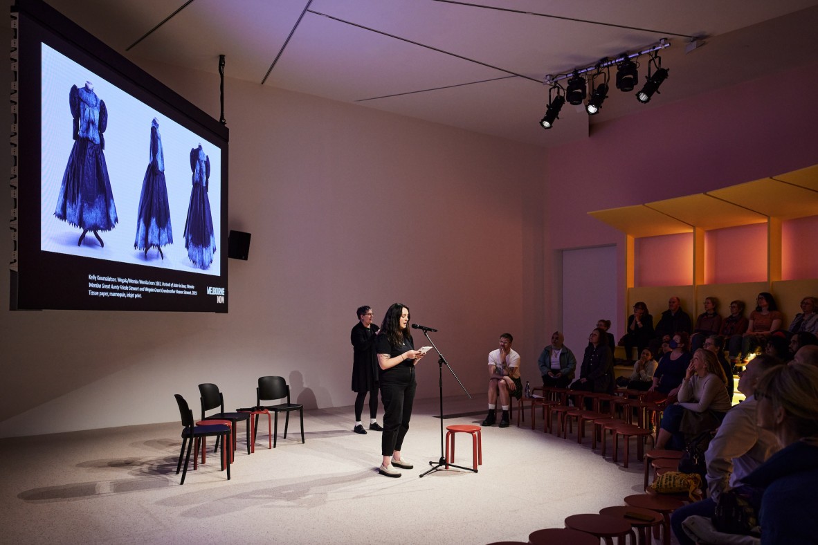 A person speaks at a microphone on a stage to a small audience; behind them is a screen showing costumes