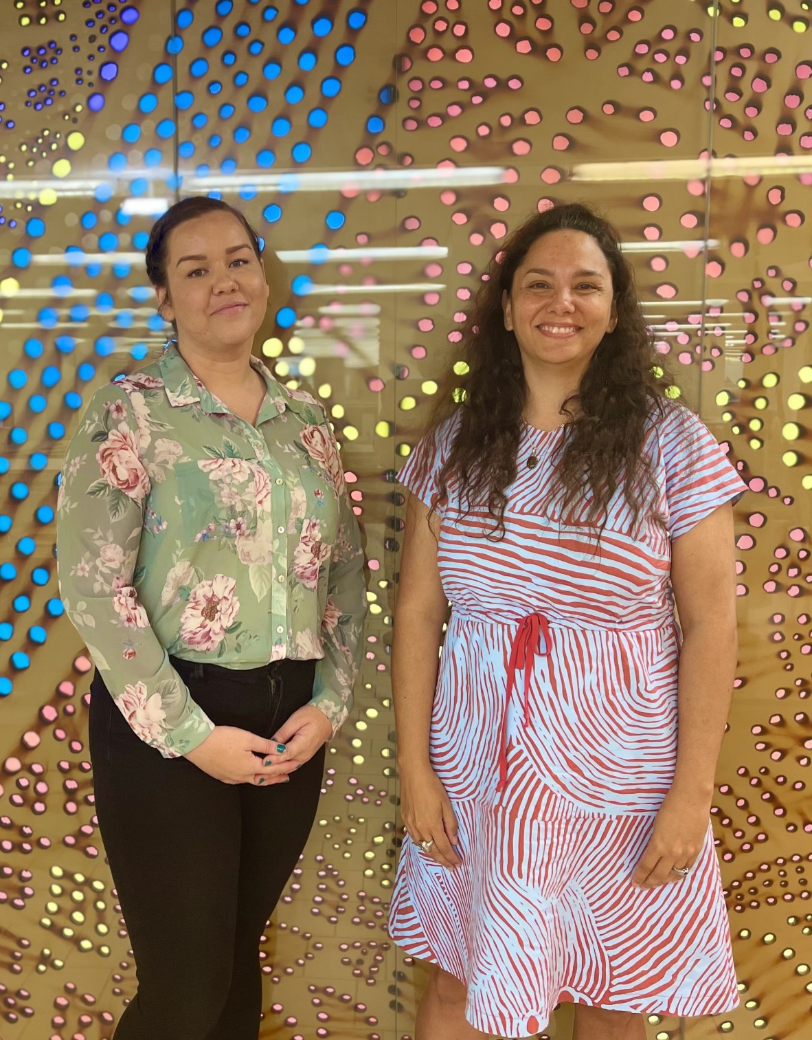 Nadia Johansen and Bianca Valentino stand smiling in front of a brown wall lit up with yellow blue and pink lights