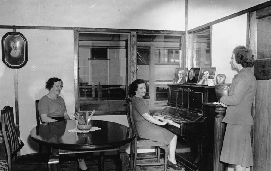 Three women in a dining room where one is playing the piano
