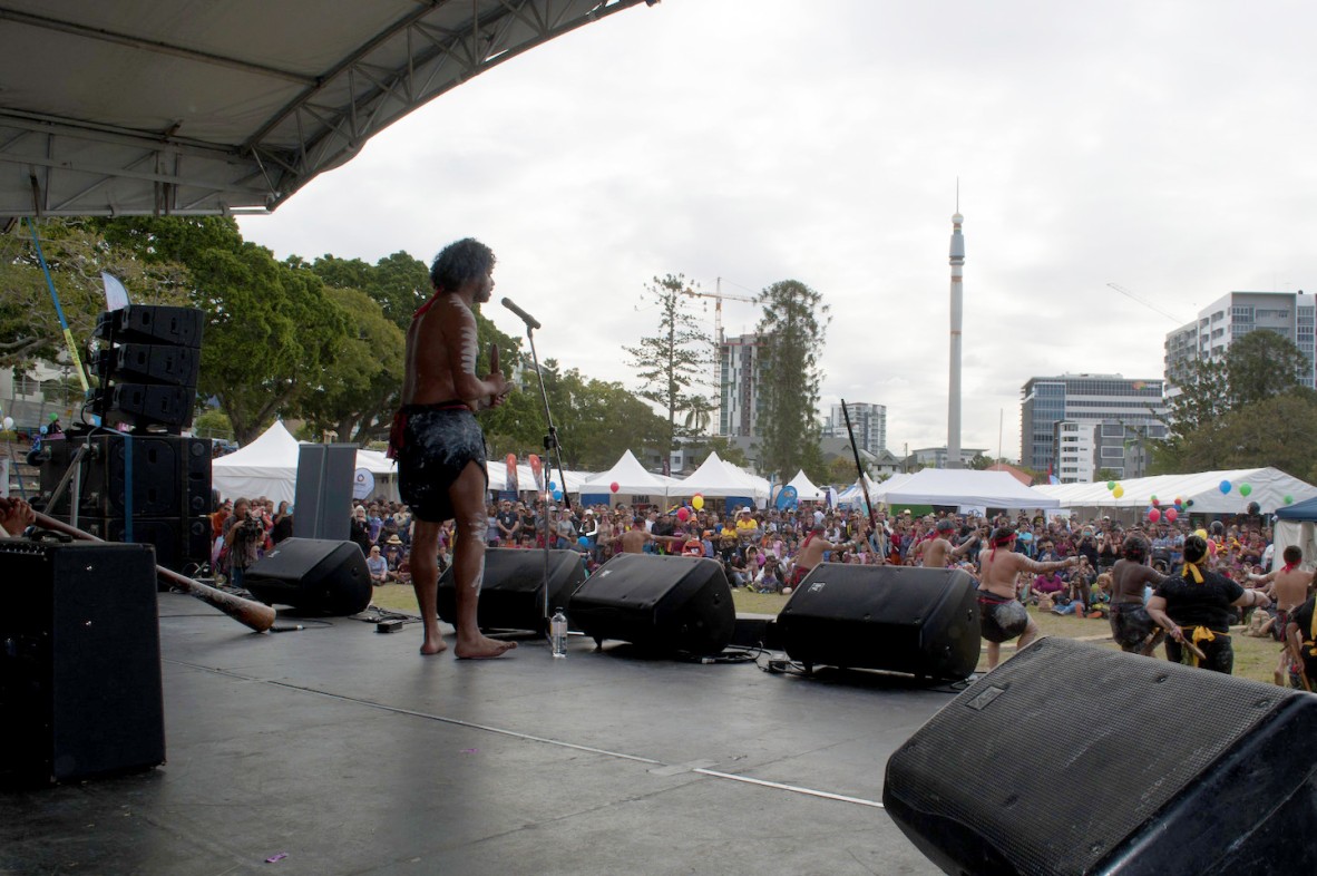 Man on stage with microphone in traditional First Nations dress. First Nations dancers are below him and a crowd watches.