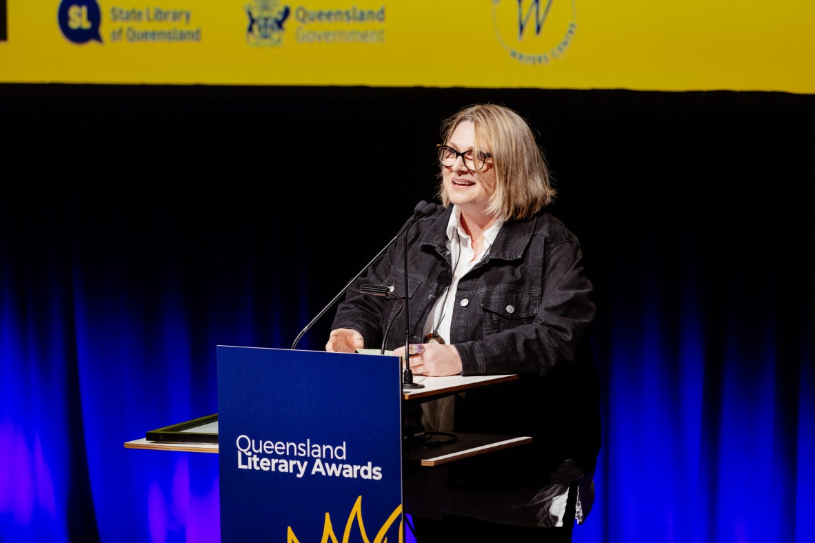 Al Campbell stands behind a lectern at the Queensland Literary Awards. She is smiling and speaking.