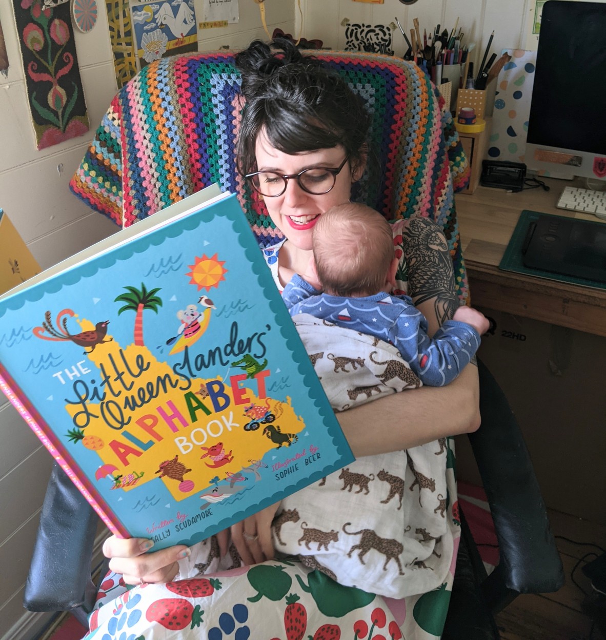 Sophie wears a bright and fruity dress and sits in a chair holding her baby and reading him The Little Queenslanders' Alphabet Book 